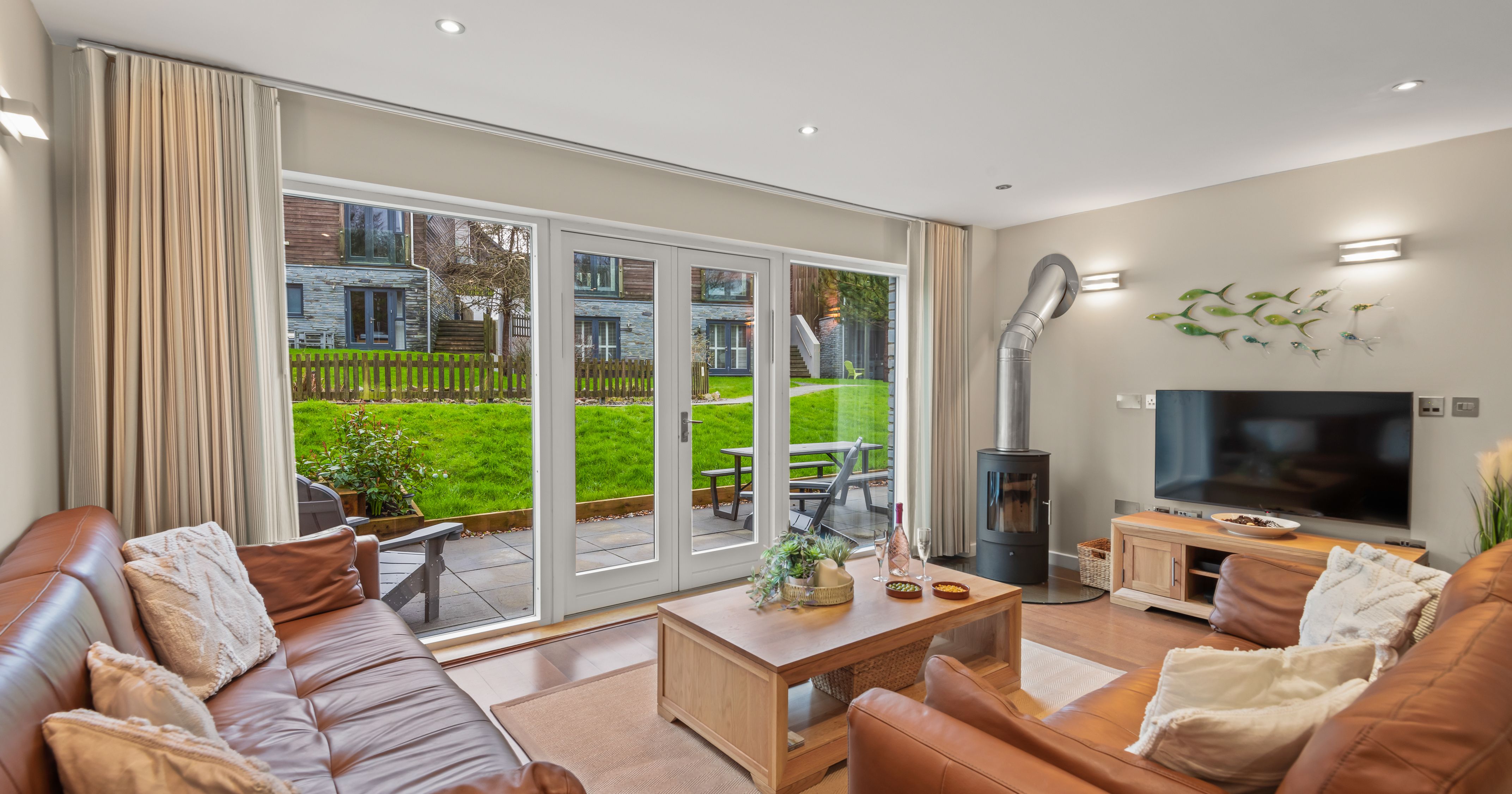 Modern living room with brown leather sofas, wood stove, TV, and large glass doors opening to a patio and green lawn.