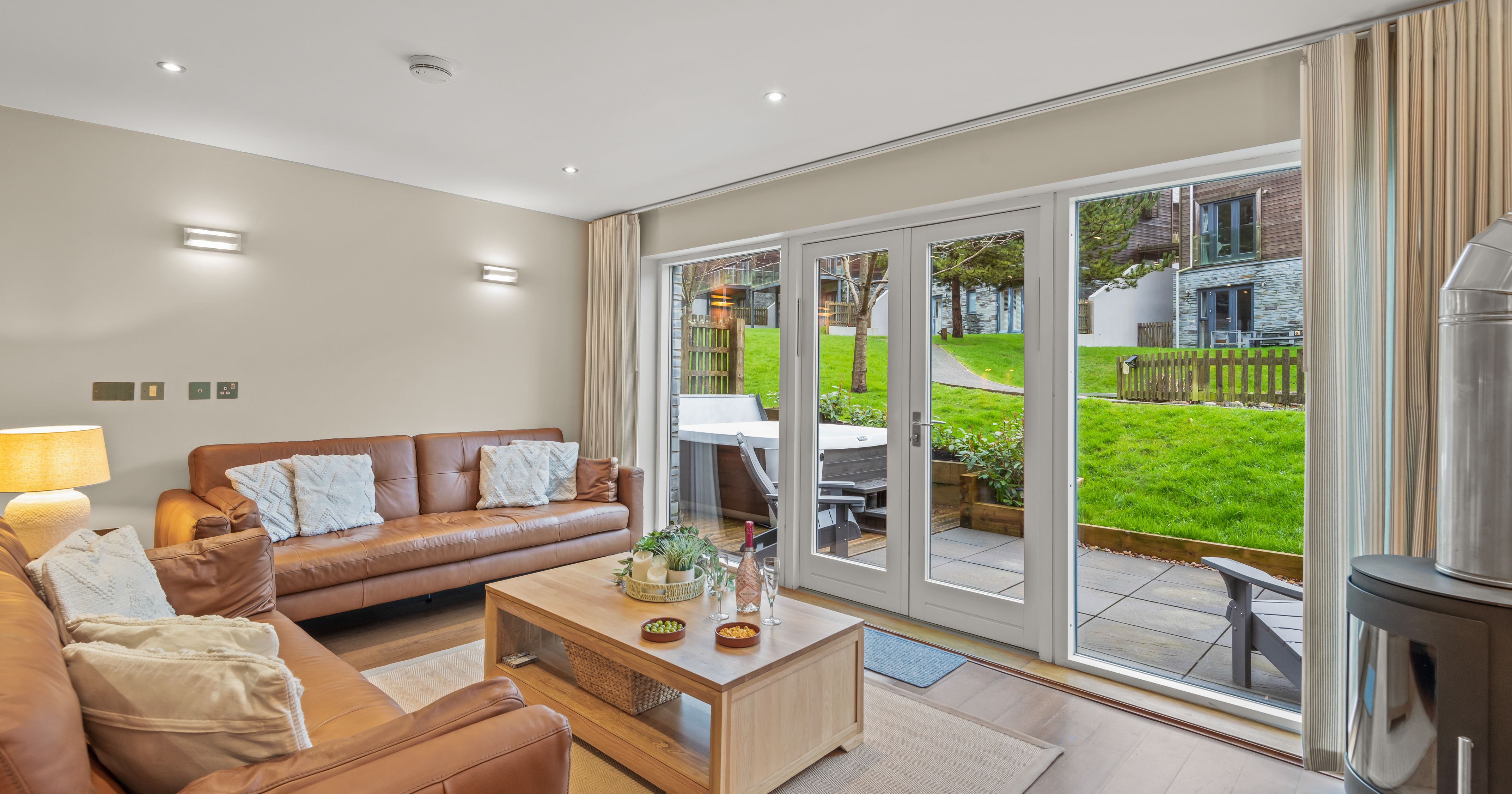 Modern living room with brown leather sofas, coffee table, and large glass doors opening to a patio and green lawn.