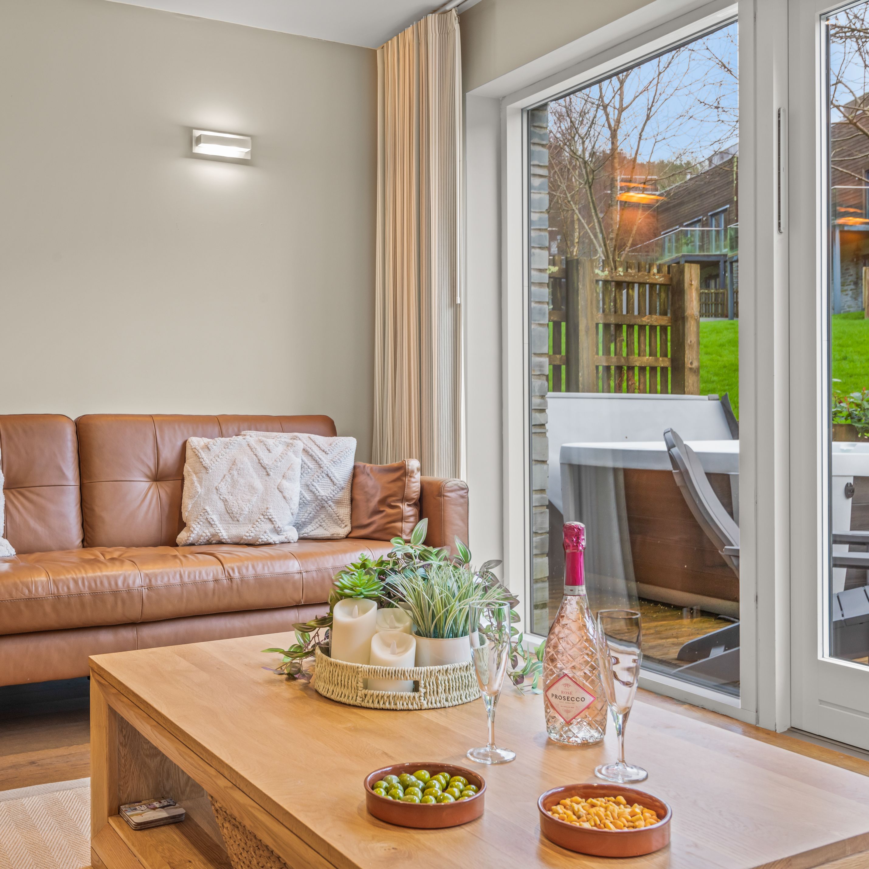 Modern living room with a brown leather sofa, large window doors, a wooden coffee table with snacks and drinks, and an outdoor hot tub view.