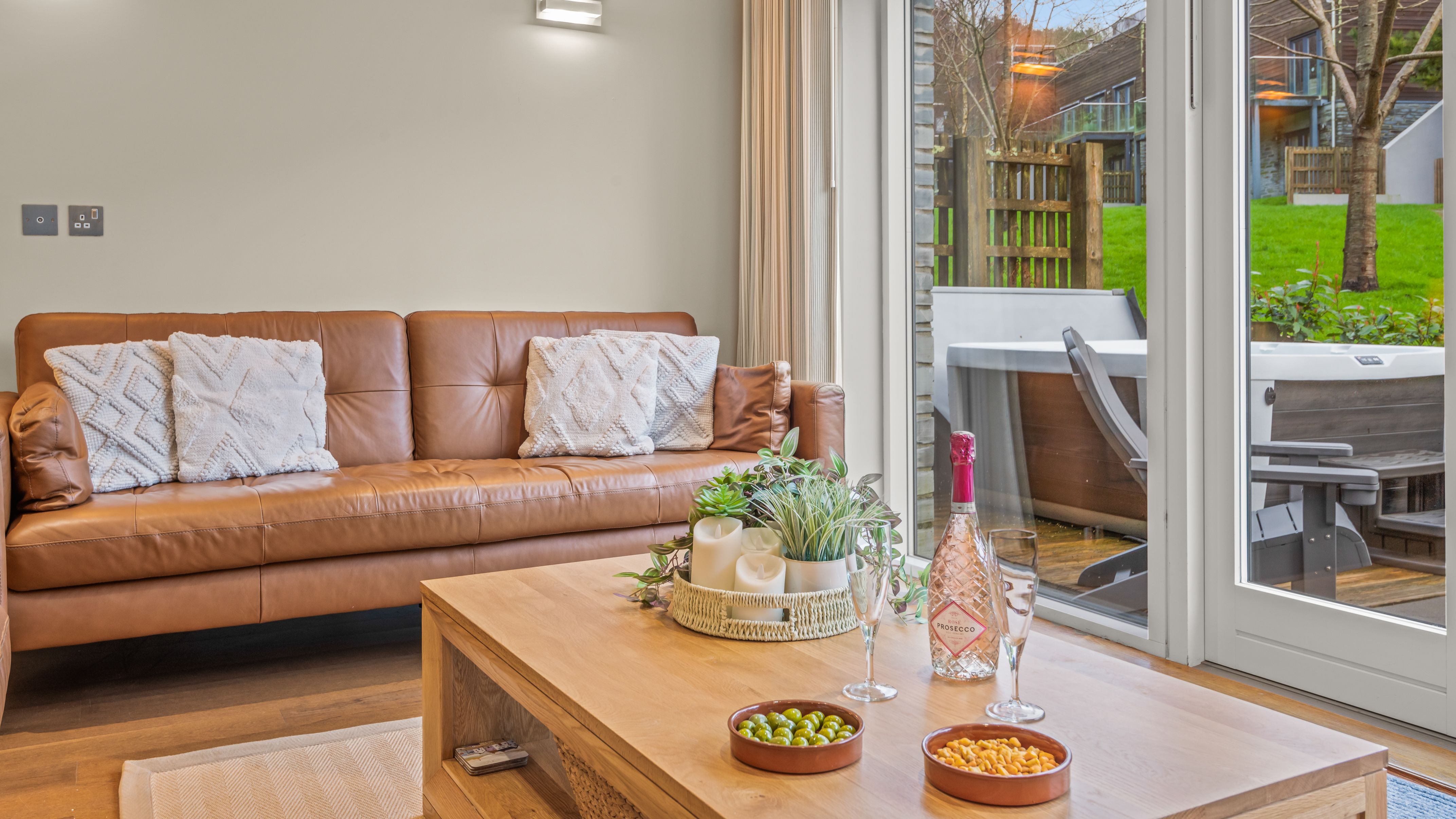 Modern living room with a brown leather sofa, large window doors, a wooden coffee table with snacks and drinks, and an outdoor hot tub view.