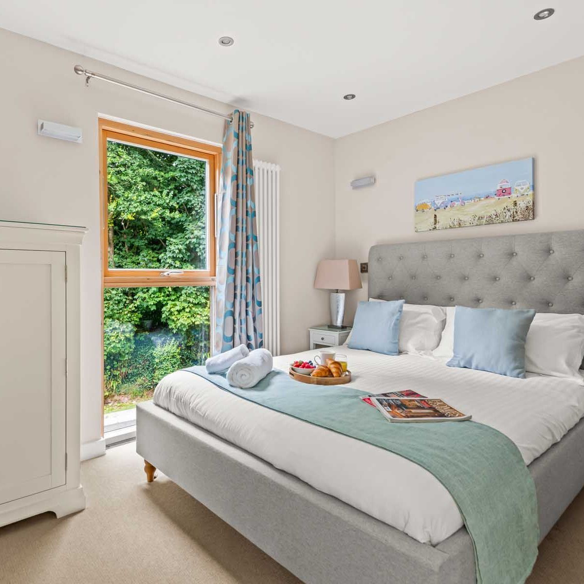 Bright and cozy bedroom with a large bed, white linens, and light blue accents, featuring a window with a view of greenery.