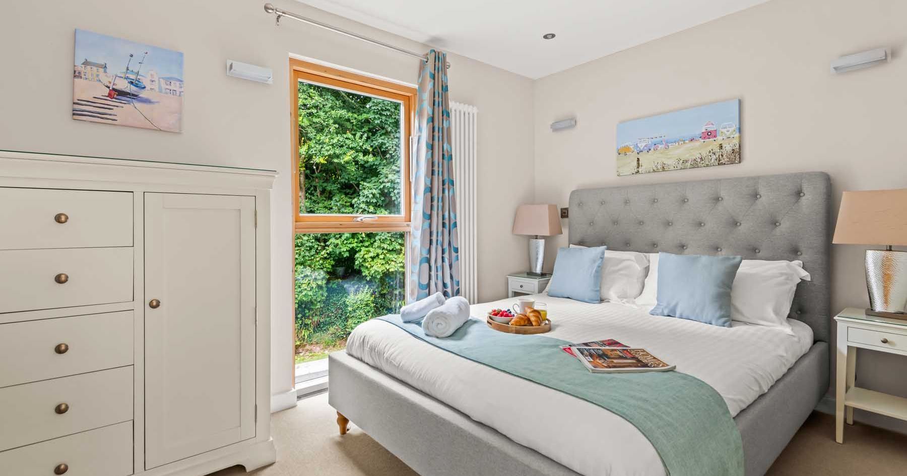 Bright and cozy bedroom with a large bed, white linens, and light blue accents, featuring a window with a view of greenery.