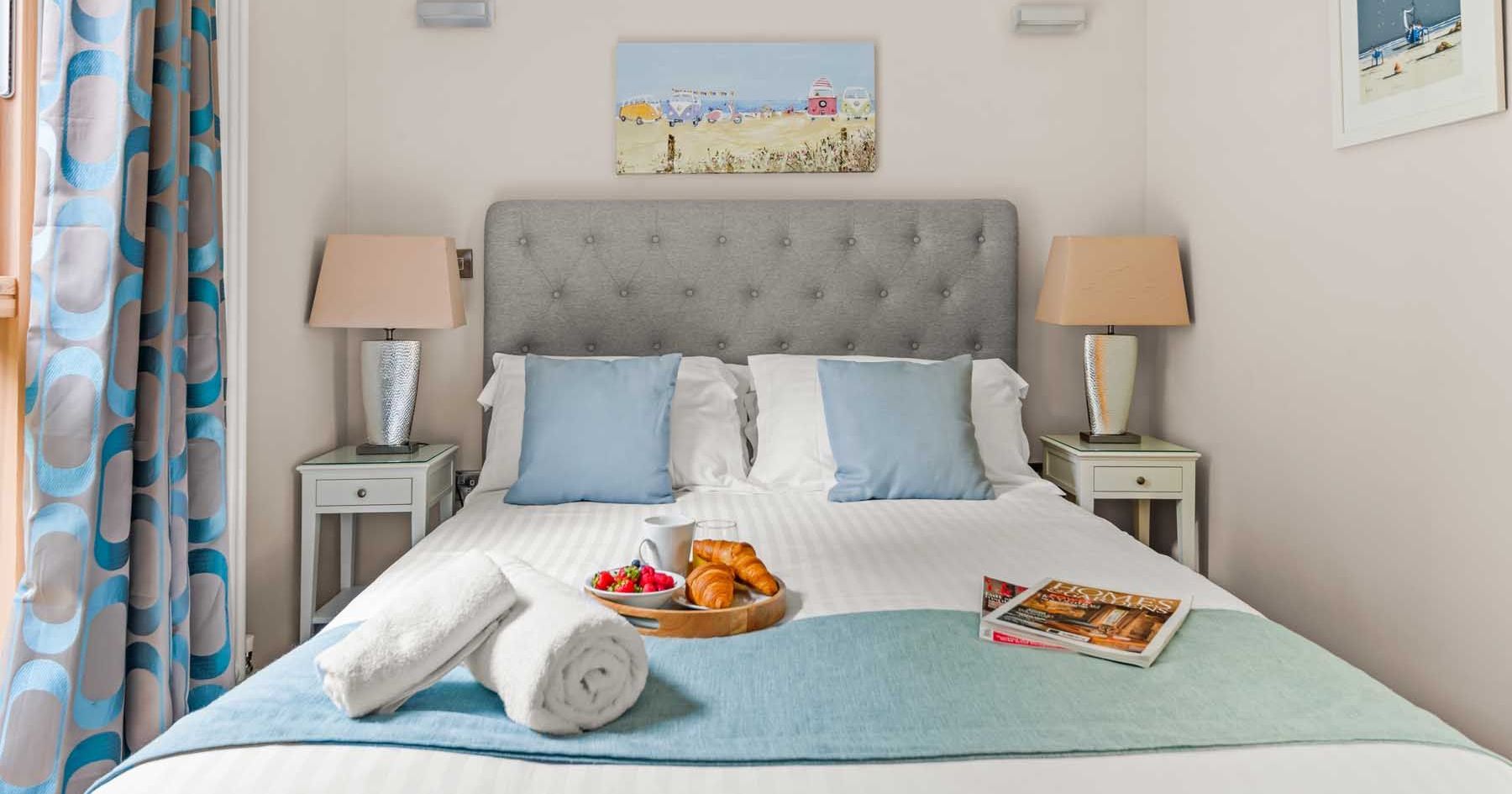 Cozy bedroom with a neatly made bed, blue accent pillows, bedside tables, lamps, and breakfast tray.