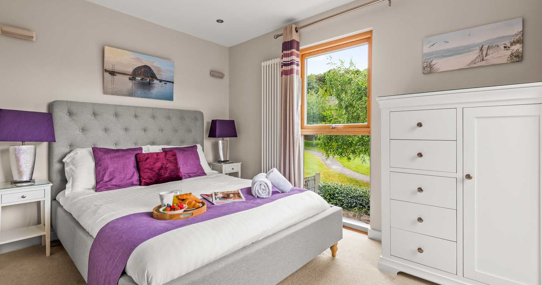 Modern bedroom with grey upholstered bed, purple accents, and large window overlooking greenery