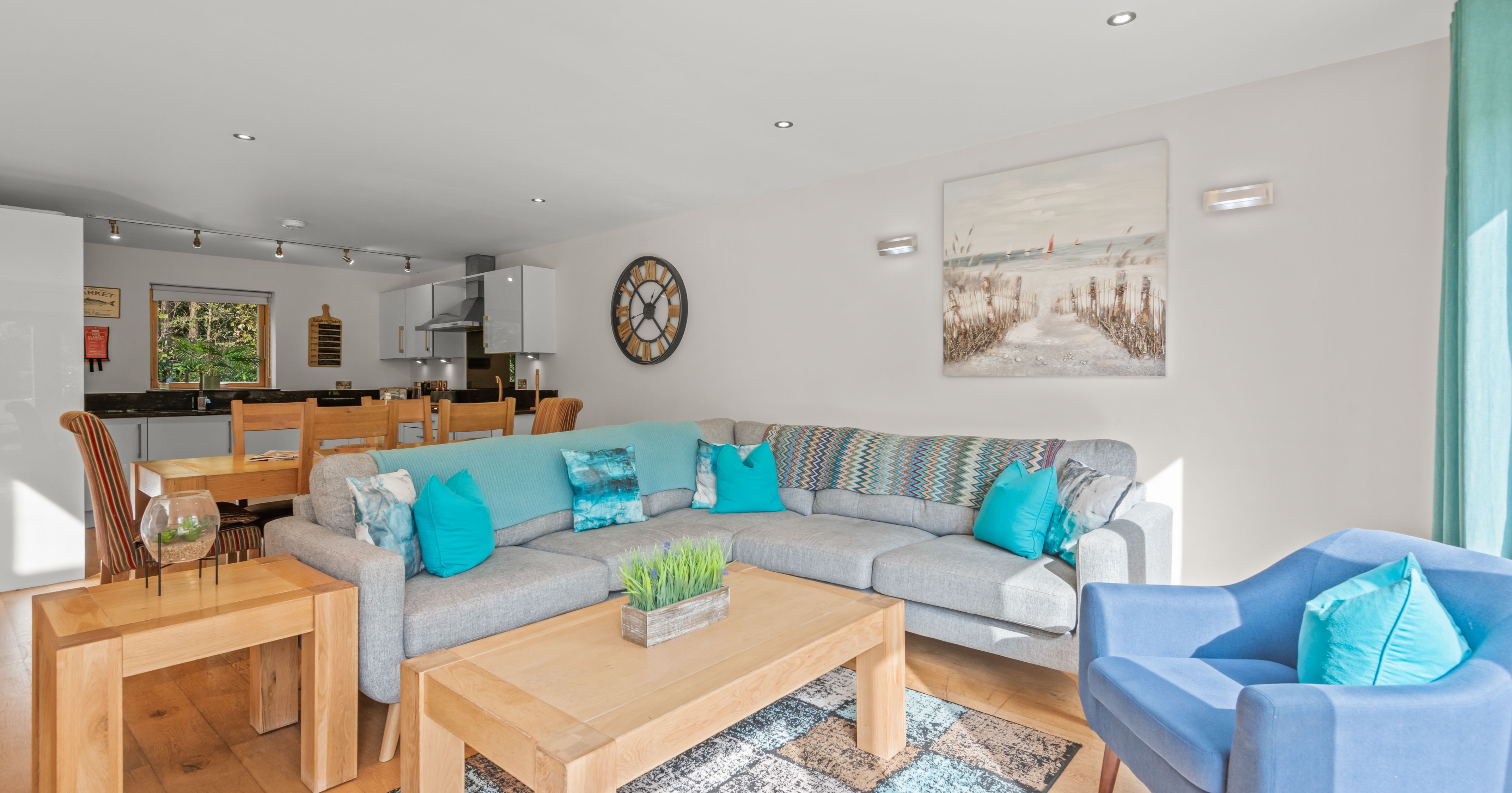 Modern open-plan living room and kitchen with light grey sectional sofa, blue accents, wooden furniture, and wall art
