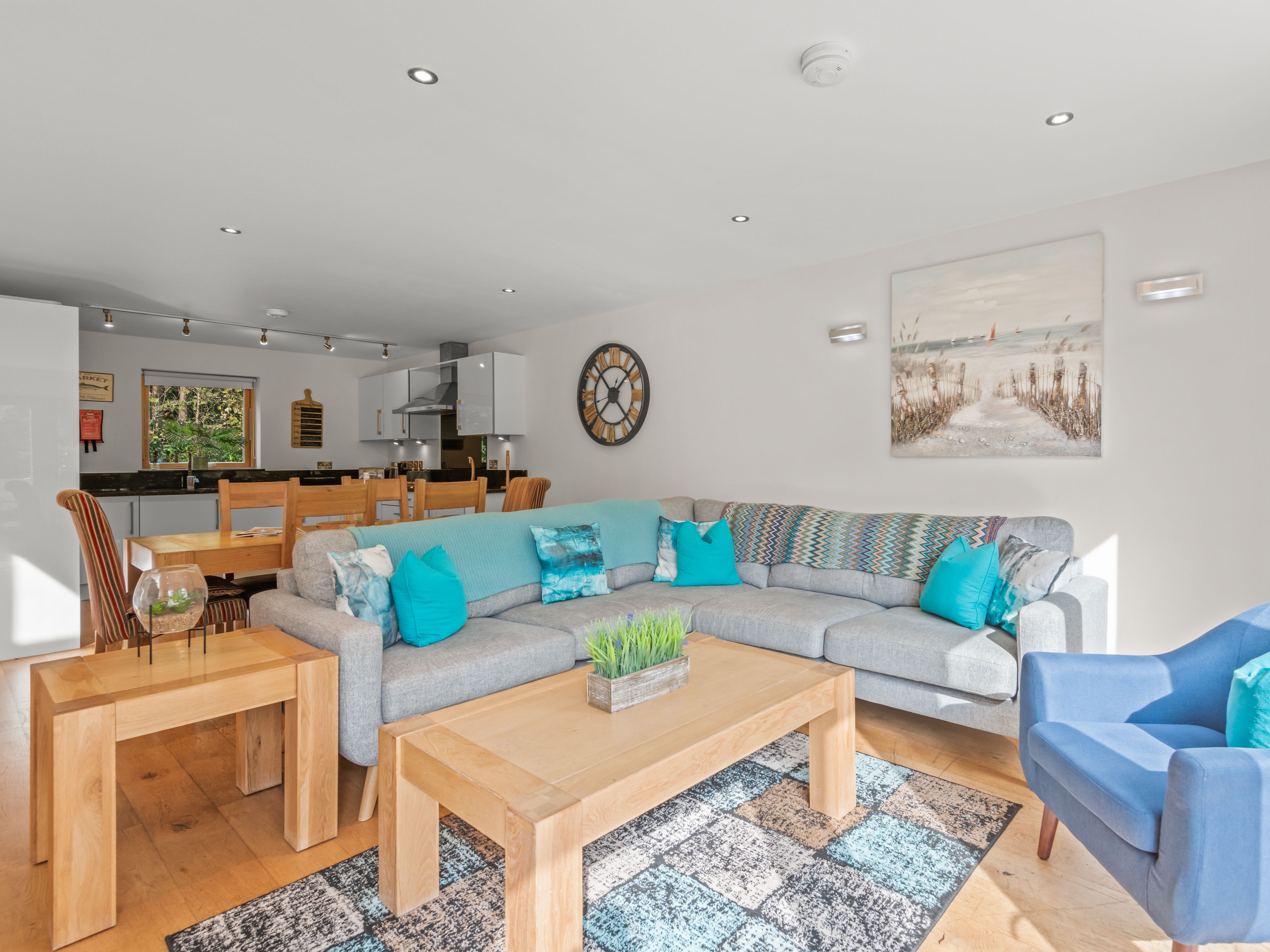 Modern open-plan living room and kitchen with light grey sectional sofa, blue accents, wooden furniture, and wall art
