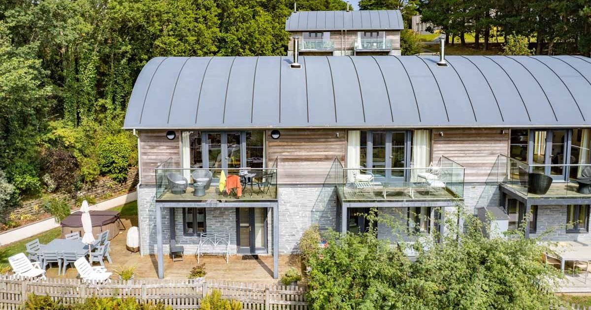 Modern house with curved metal roof, glass balconies, and patio in a wooded setting