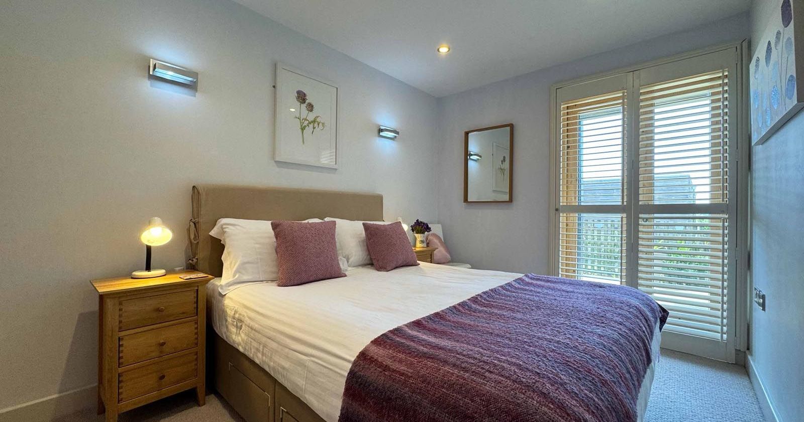 Bright modern bedroom with a double bed, wooden nightstand, and large window with blinds.