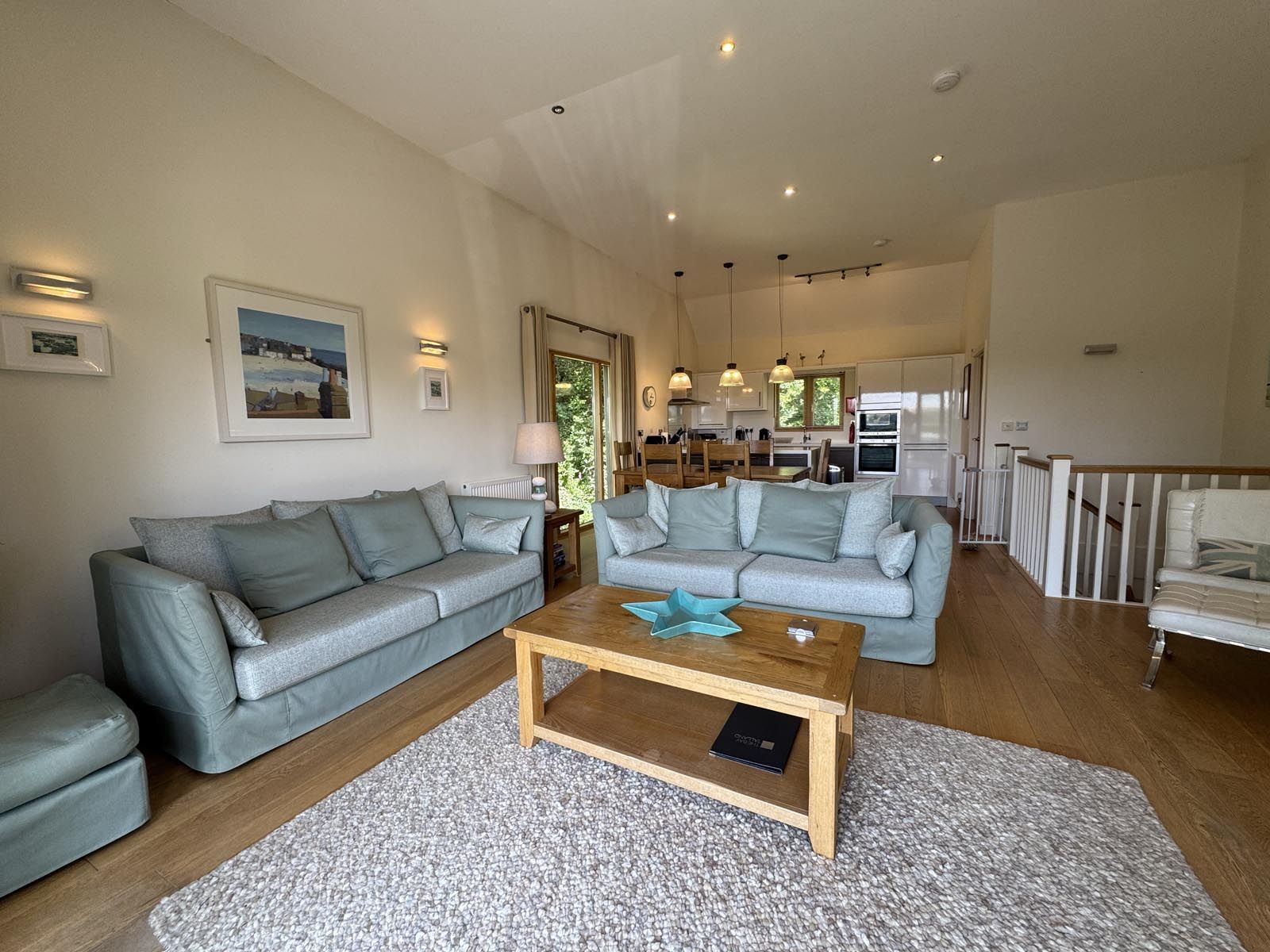 Modern, open-plan living room and kitchen with light blue sofas, wooden coffee table, and neutral decor.
