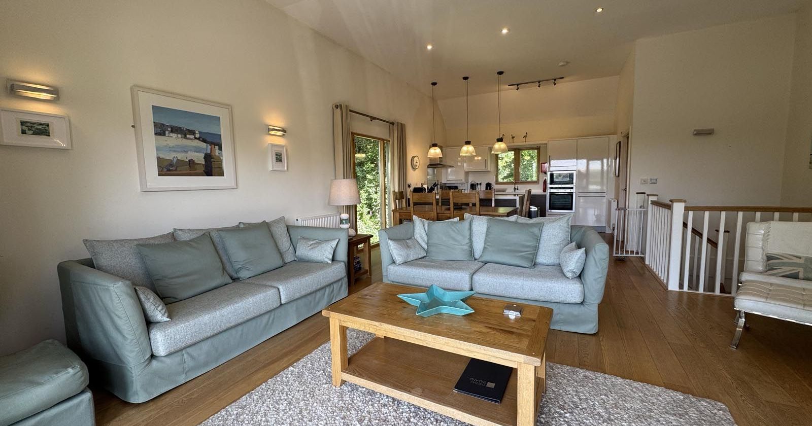 Modern, open-plan living room and kitchen with light blue sofas, wooden coffee table, and neutral decor.