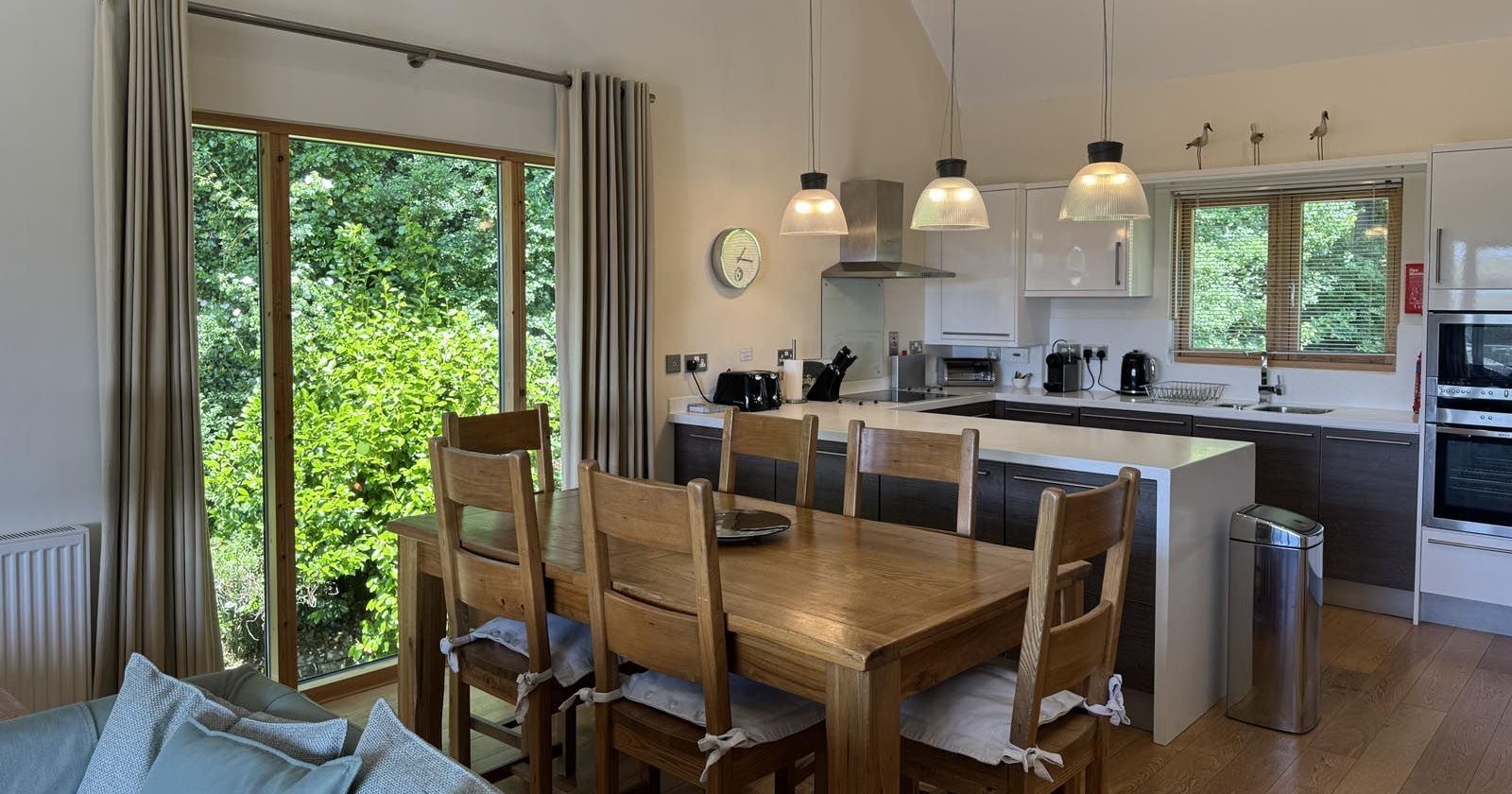 Modern open-plan kitchen and dining area with wooden furniture and large window view of greenery.