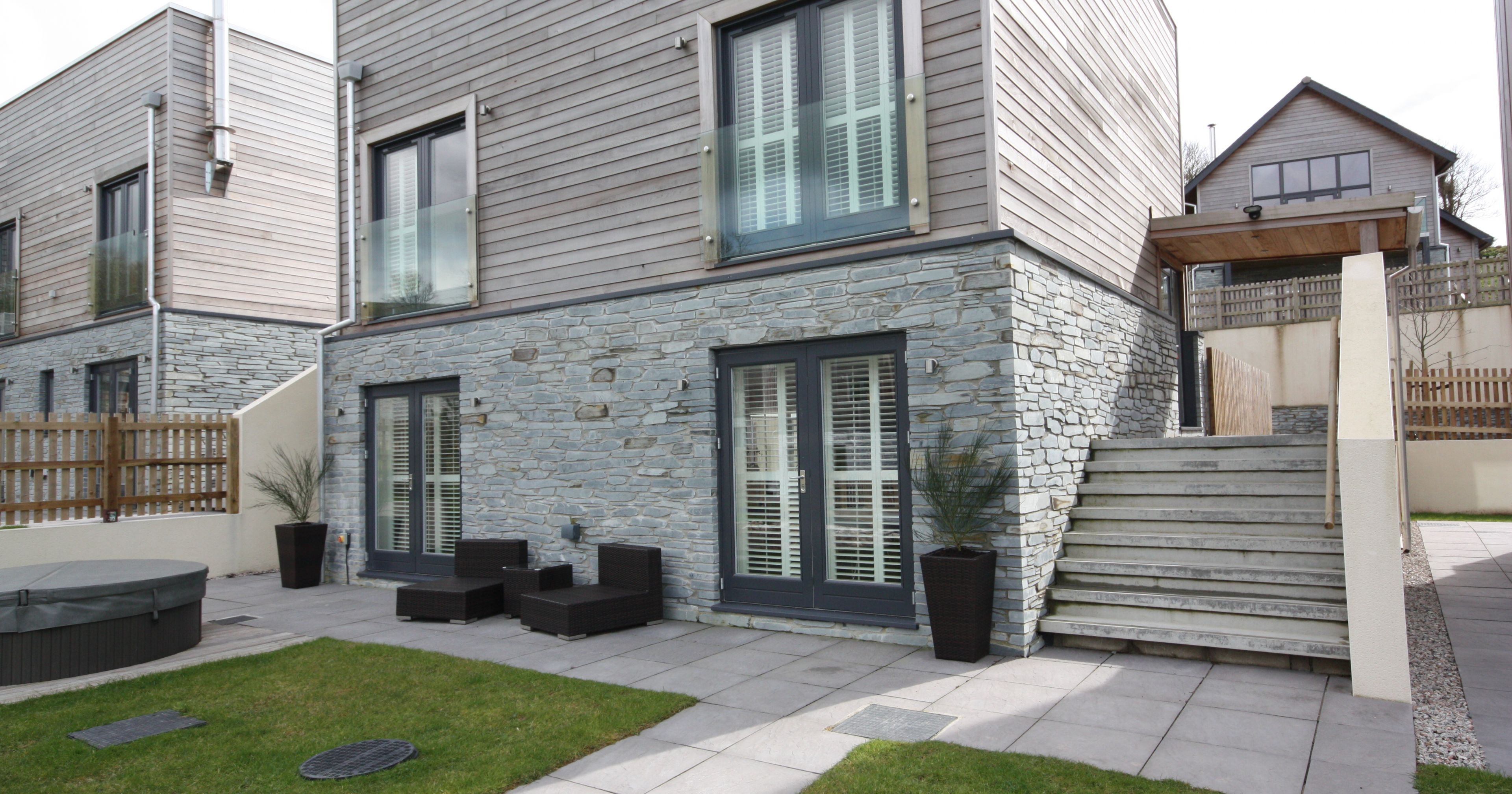 Modern two-story house with stone and wood exterior, large windows with shutters, outdoor seating area, hot tub, and steps leading to an upper level.