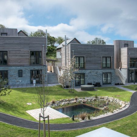 Modern two-story buildings with wooden and stone facades, surrounded by green lawns, a small pond, paths, and young trees under a partly cloudy sky.