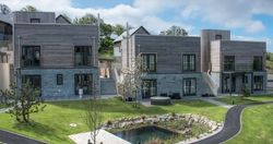 Modern two-story buildings with wooden and stone facades, surrounded by green lawns, a small pond, paths, and young trees under a partly cloudy sky.