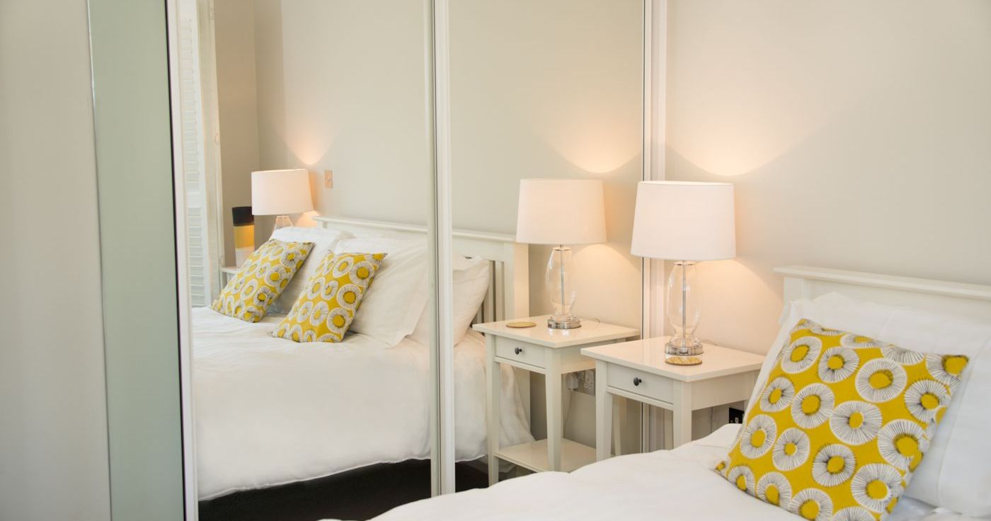 Modern bedroom with white decor, yellow patterned pillows, mirrored wardrobe, and bedside lamps