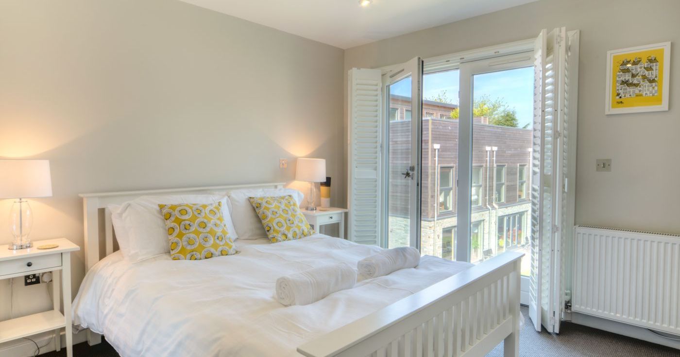 Bright modern bedroom with double bed, white linens, and open glass doors to a balcony.