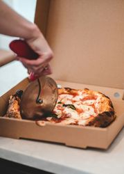 Hand using a pizza cutter to slice a pizza in a cardboard box