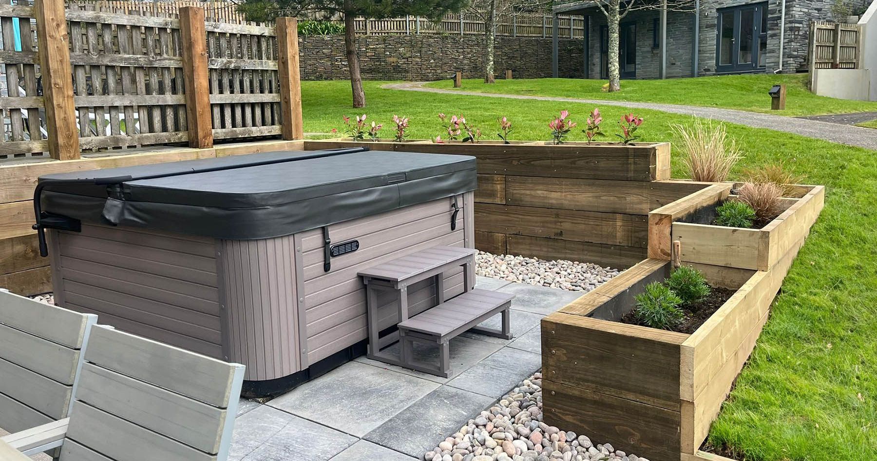 Modern backyard with a covered hot tub, stone patio, wooden raised garden beds, and green lawn.