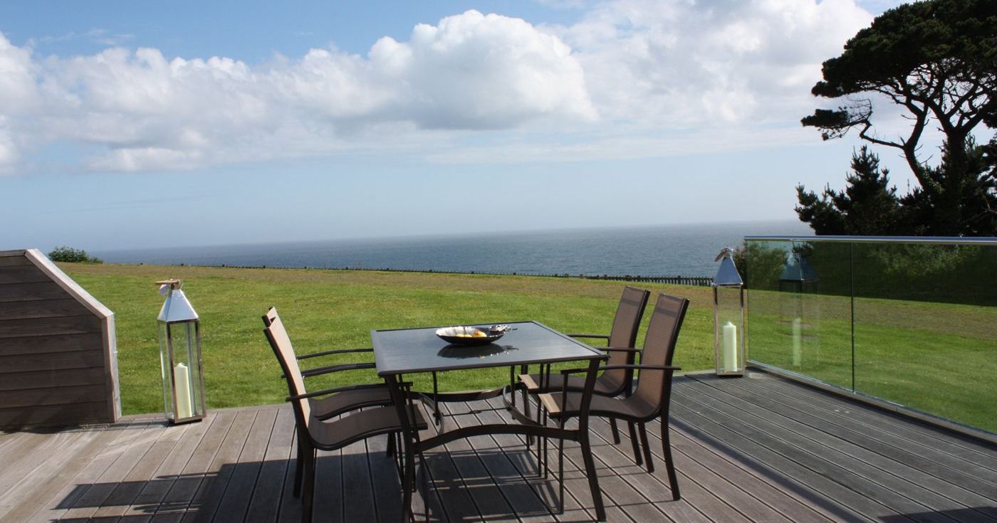 Outdoor patio with table and chairs on a wooden deck overlooking a grassy lawn and the ocean, with glass railing and large lanterns.