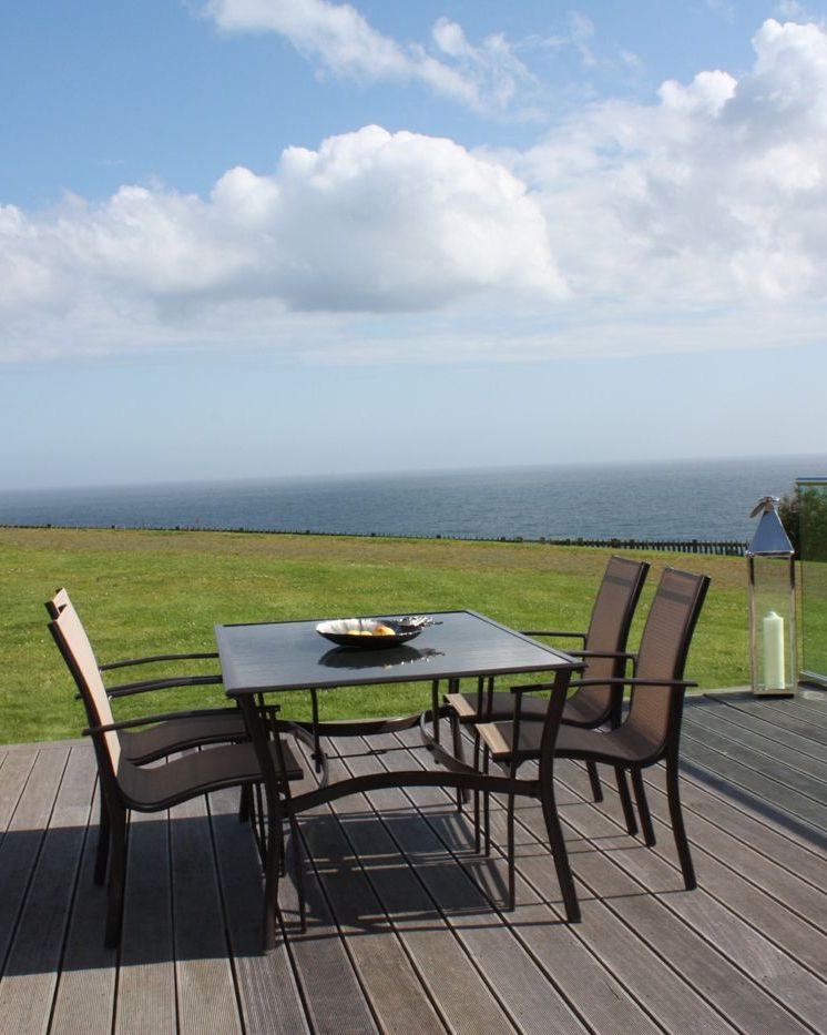 Outdoor patio with table and chairs on a wooden deck overlooking a grassy lawn and the ocean, with glass railing and large lanterns.