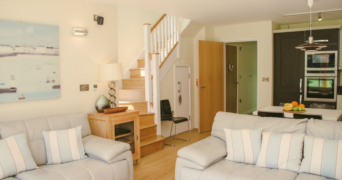 Modern living room and kitchen area with light-colored sofas, a wooden staircase, and open kitchen.