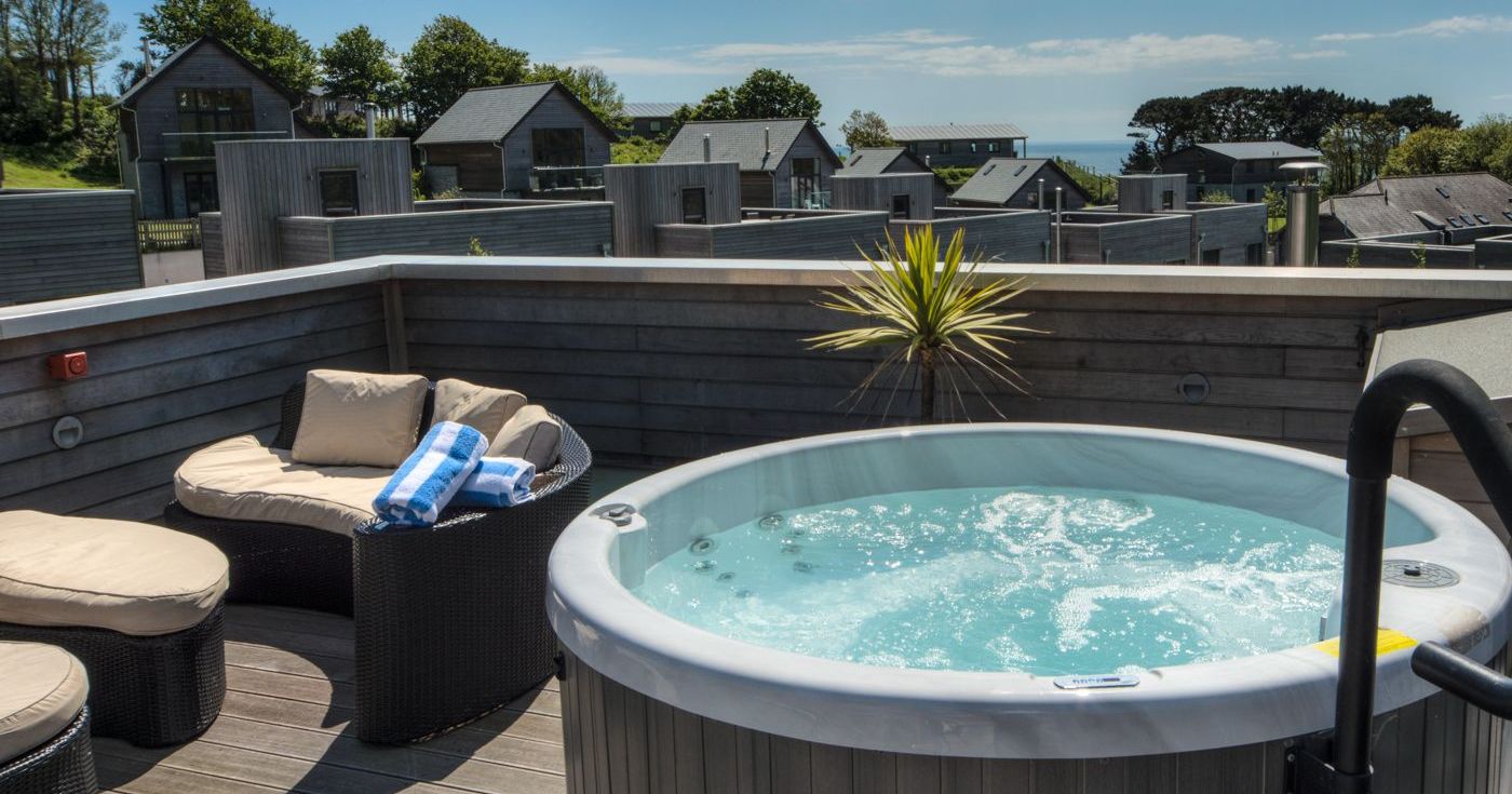 Outdoor hot tub on a balcony with seating and a view of modern houses and trees under a blue sky.