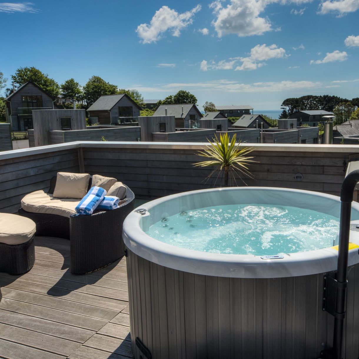 Outdoor hot tub on a balcony with seating and a view of modern houses and trees under a blue sky.