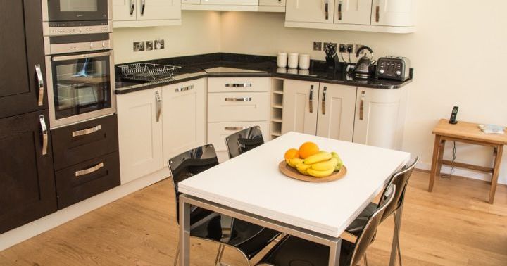 Modern kitchen with white cabinets, black countertops, a wooden table with fruit, and black chairs.