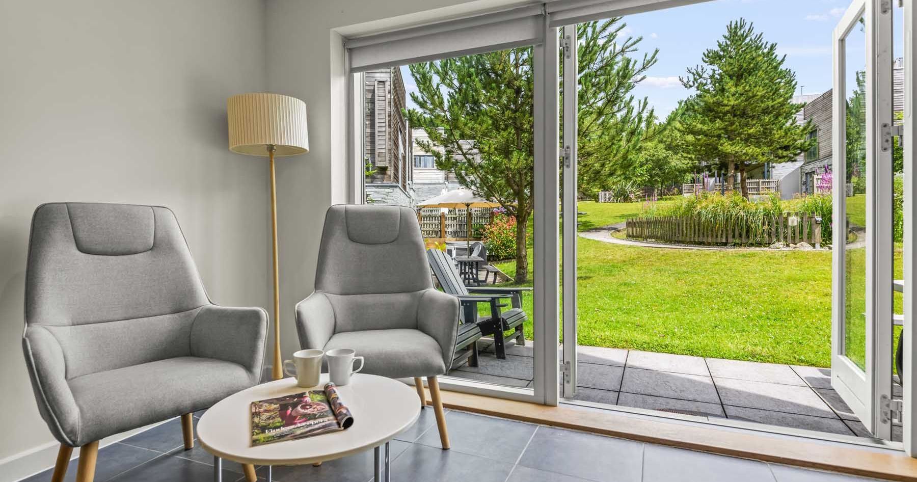 Modern sitting area with two gray chairs, a floor lamp, a small table with cups and a magazine, and open doors leading to a lush garden.