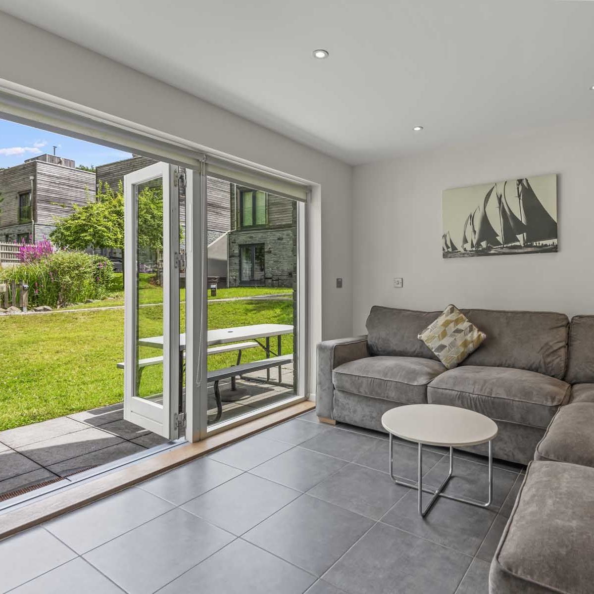 Modern living room with grey sectional sofa, open patio doors, and view of garden and contemporary buildings outside.