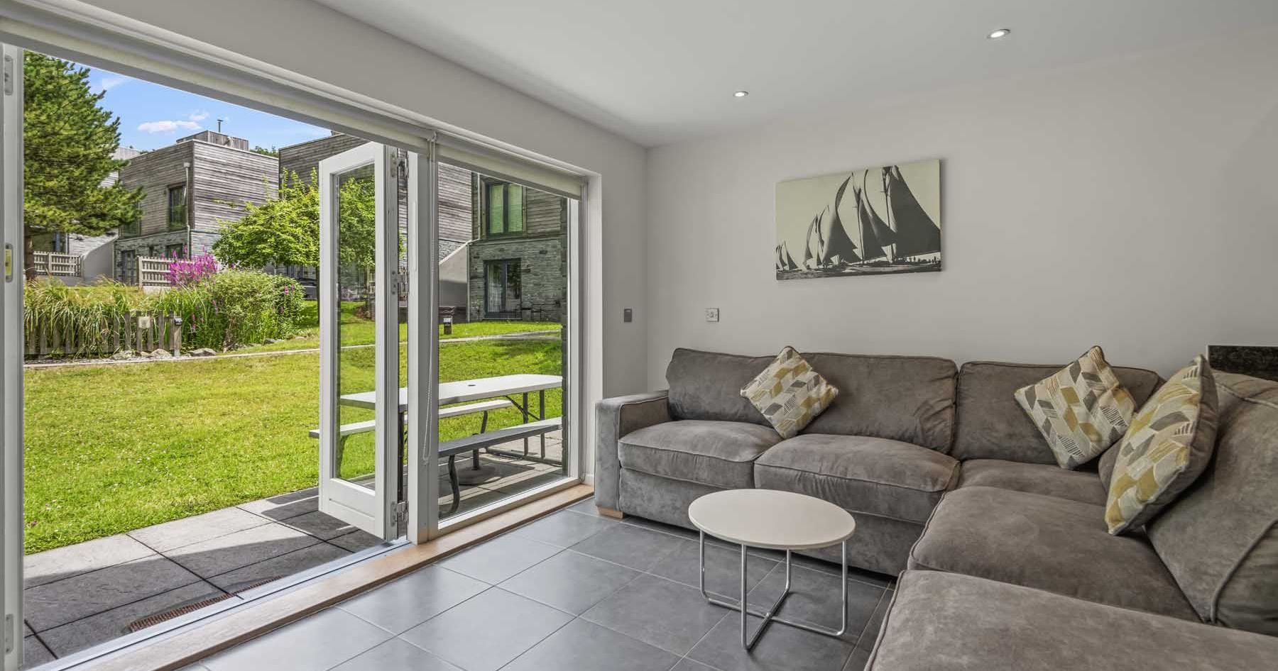 Modern living room with grey sectional sofa, open patio doors, and view of garden and contemporary buildings outside.