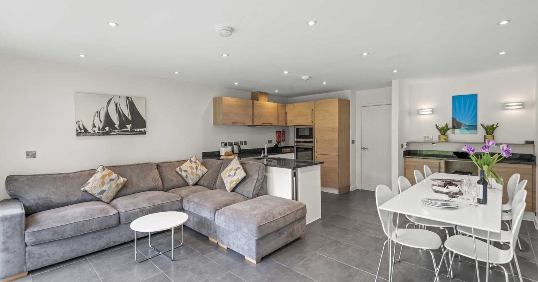 Modern open-plan living room and kitchen with grey sofa, wooden cabinets, and dining table
