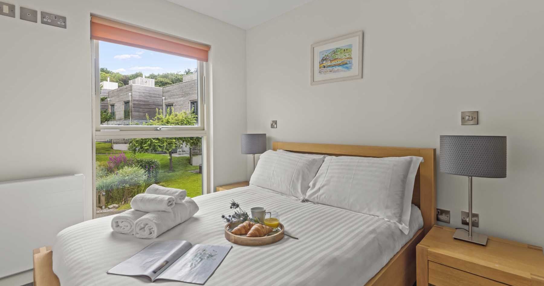 Modern bedroom with neatly made bed, breakfast tray, and window view of garden and buildings.