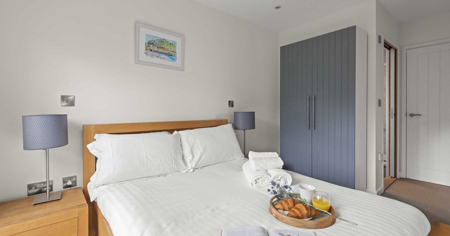 Modern bedroom with neatly made bed, blue wardrobe, bedside tables with lamps, and breakfast tray with croissants and juice.