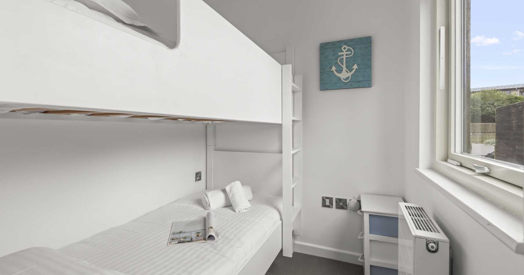 Small bedroom with white bunk beds, a window, and a nautical-themed anchor artwork.