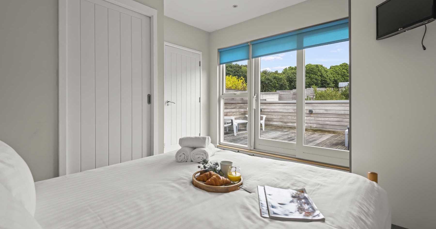 Modern bedroom with white decor, a tray with breakfast, and a view of a wooden deck through large glass doors.