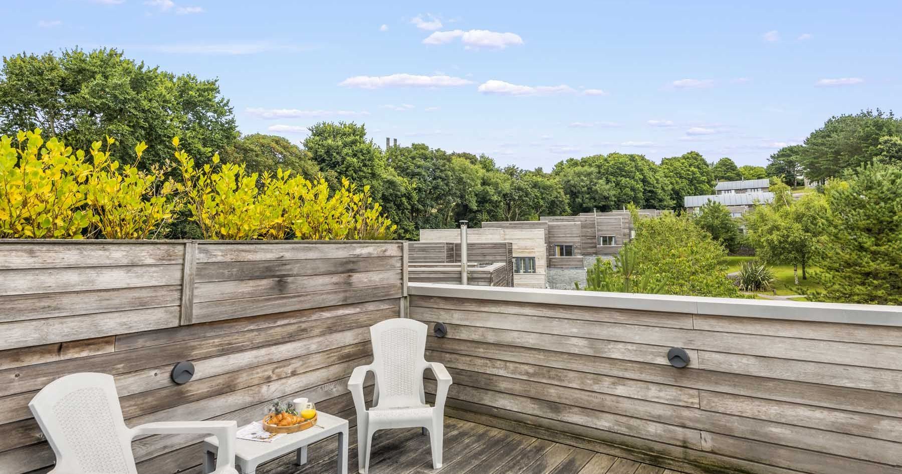 Wooden rooftop terrace with two white chairs and a small table overlooking greenery and modern buildings.