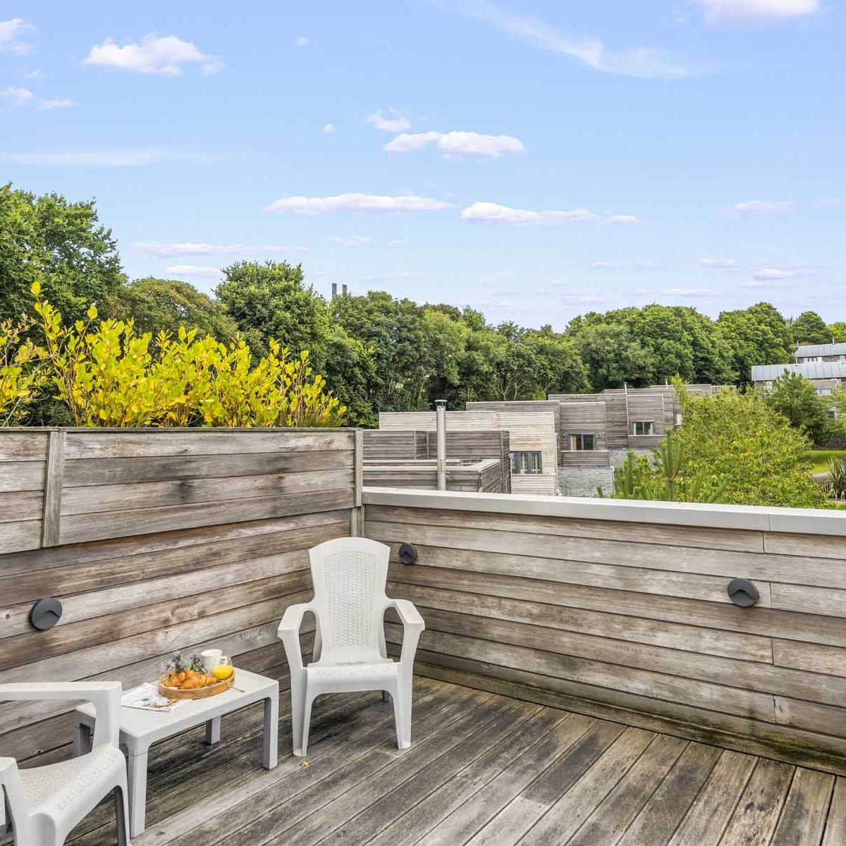 Wooden rooftop terrace with two white chairs and a small table overlooking greenery and modern buildings.