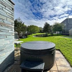 Covered hot tub in a grassy backyard with modern buildings and trees under a partly cloudy sky.