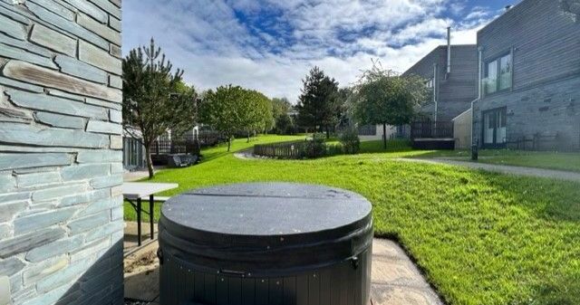 Covered hot tub in a grassy backyard with modern buildings and trees under a partly cloudy sky.