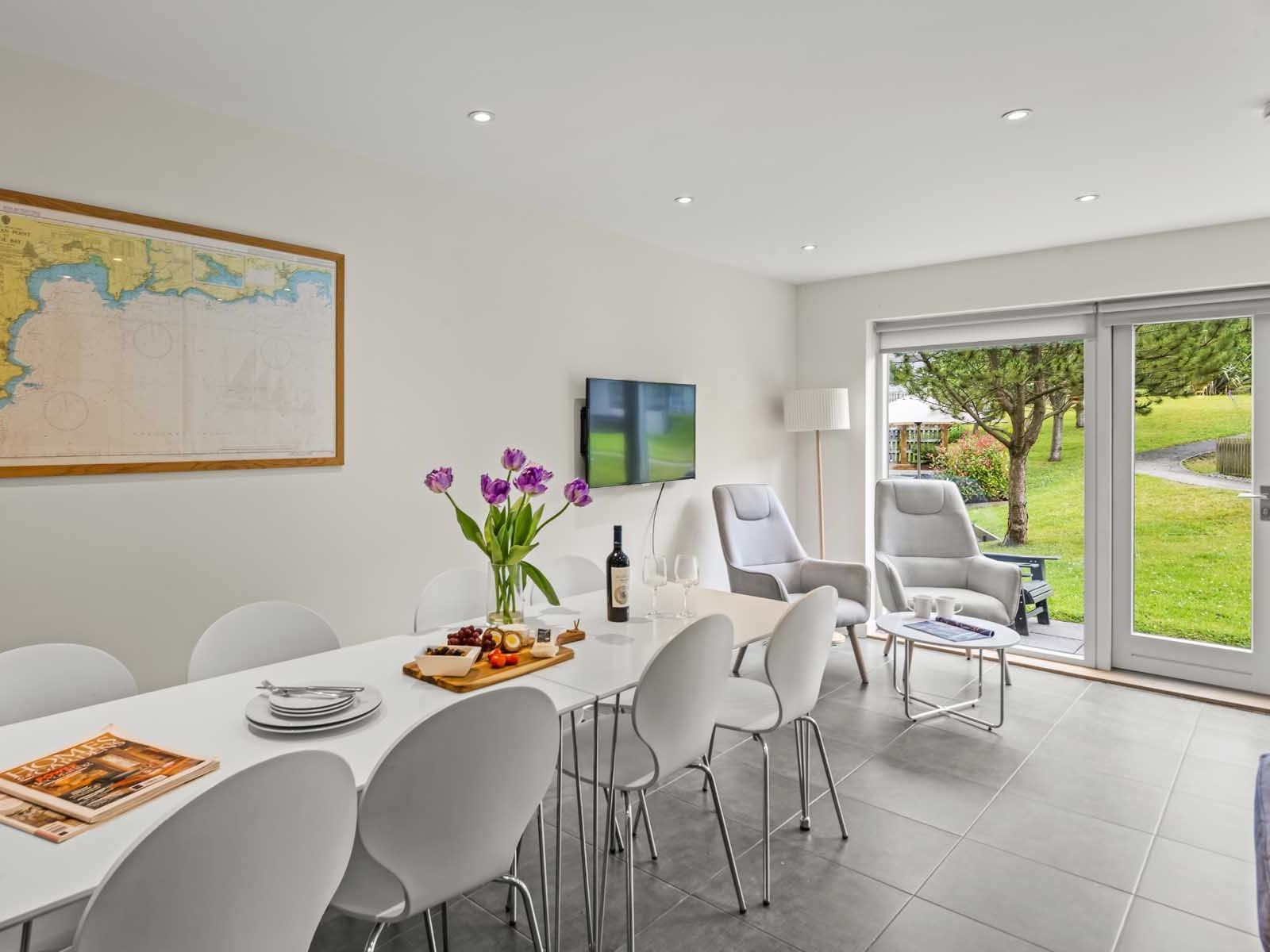 Modern dining and living room with white furniture, wall art, and large windows overlooking a green garden