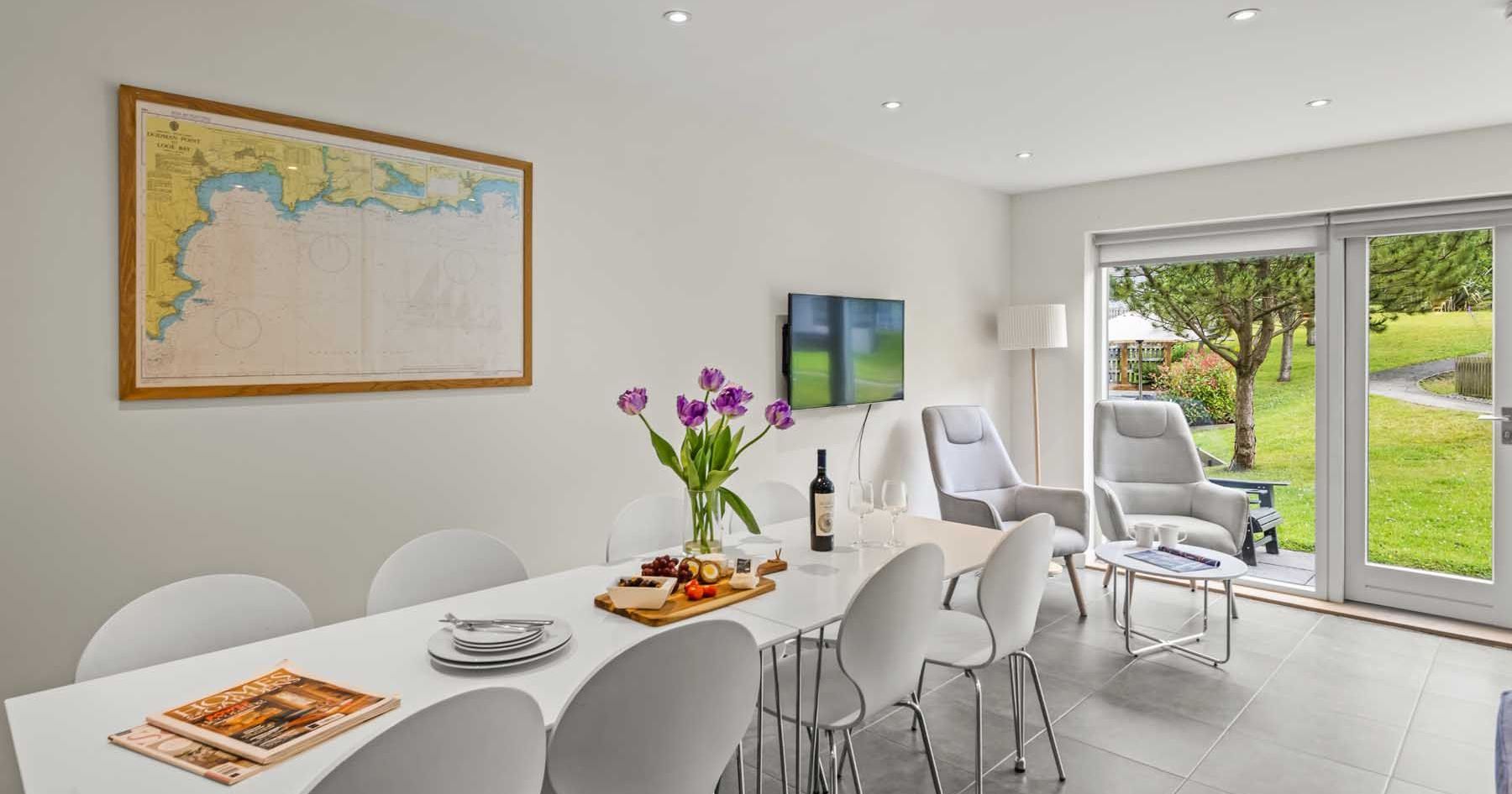 Modern dining and living room with white furniture, wall art, and large windows overlooking a green garden