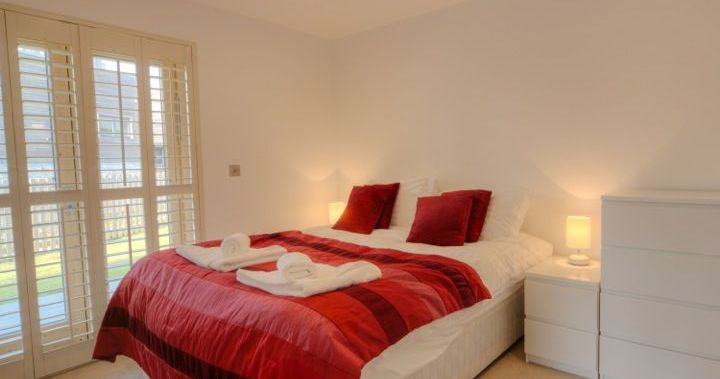 Modern bedroom with a double bed covered in a red duvet and pillows, white dresser and nightstand, and large windows with shutters.
