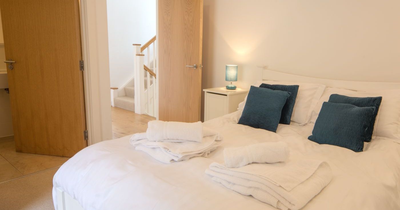 Bright bedroom with neatly made bed, teal pillows, and folded white towels. Two open wooden doors reveal a bathroom and staircase.