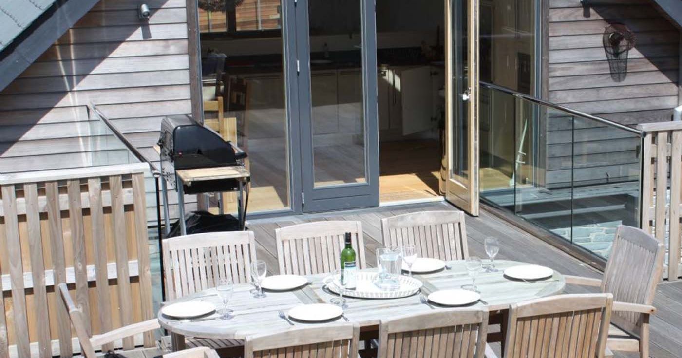 Outdoor patio with wooden dining table and chairs, set for a meal, adjacent to house with glass doors and a grill.