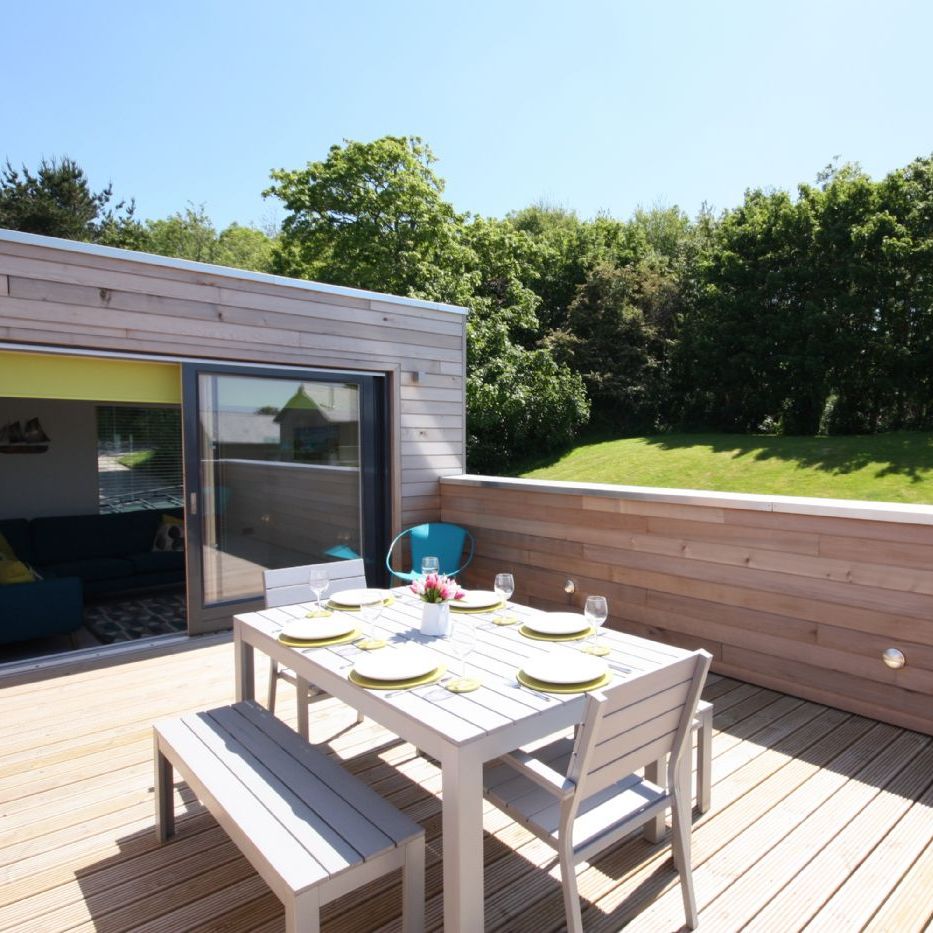 Modern outdoor wooden deck with dining table and benches, surrounded by greenery.