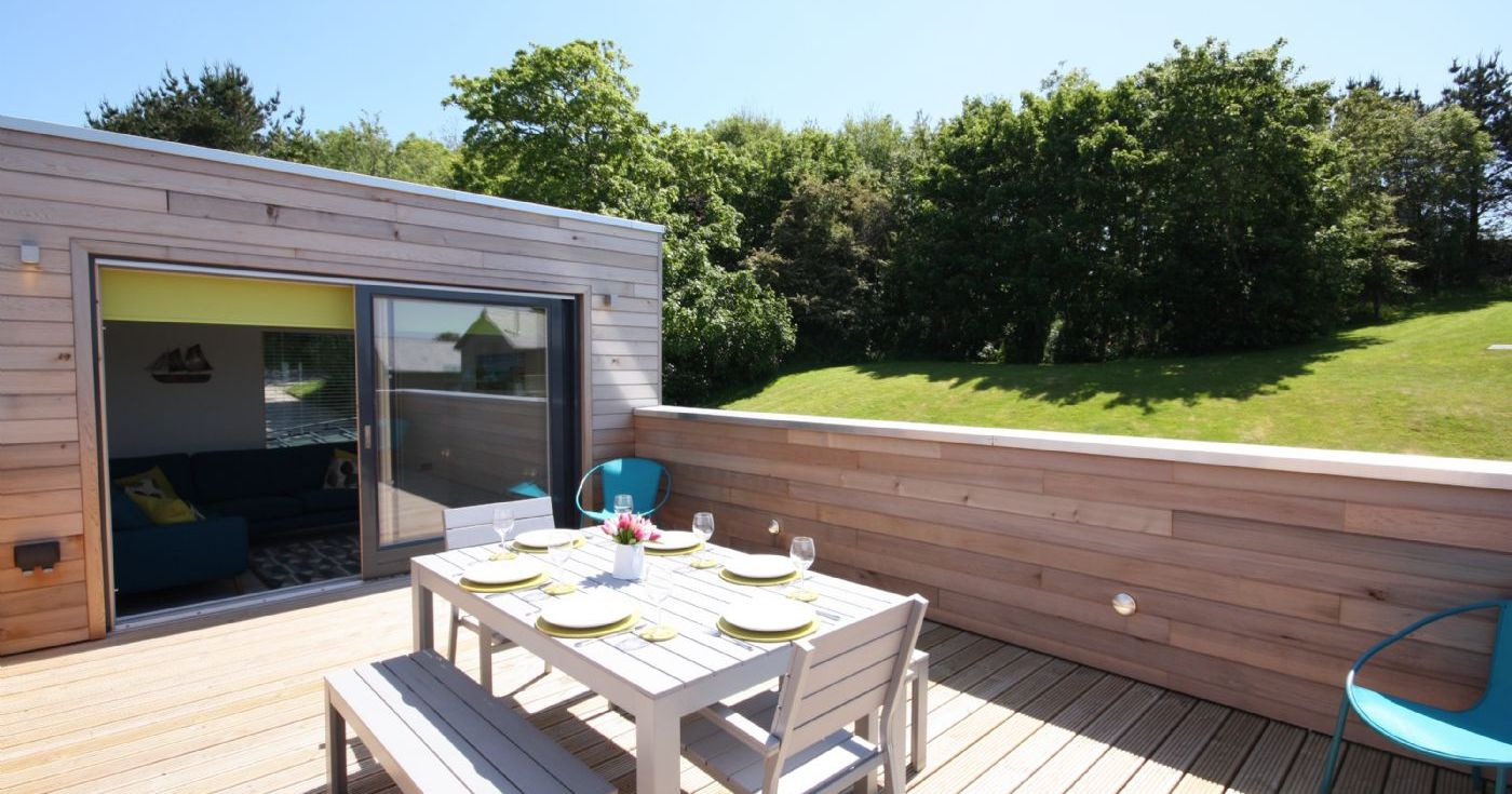 Modern outdoor wooden deck with dining table and benches, surrounded by greenery.