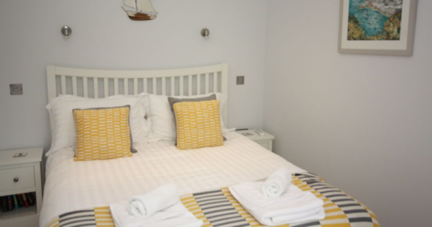 Neatly made bed with white and yellow bedding and two rolled towels, yellow and gray accent pillows, nautical wall decor, and a framed picture.