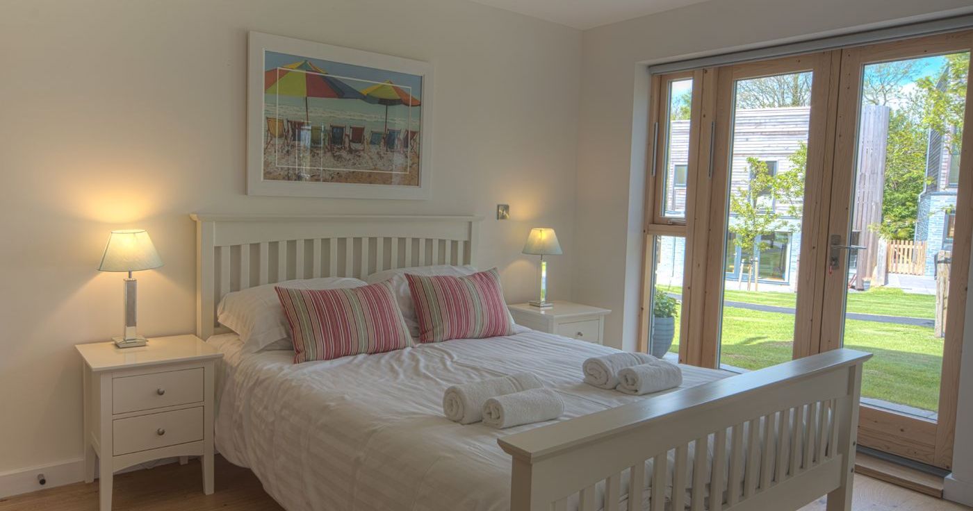 Bright, modern bedroom with a white wooden bed, striped pillows, bedside tables, and large windows overlooking a green garden.