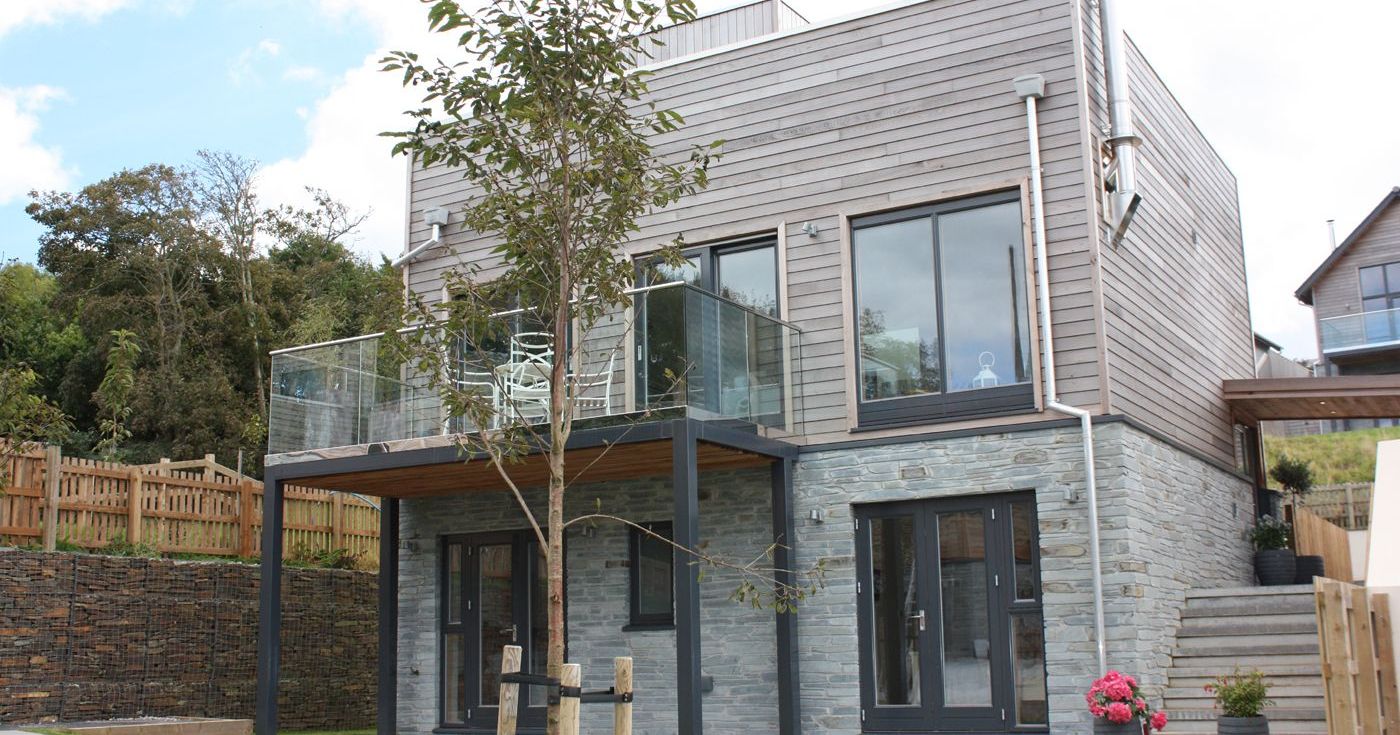 Modern two-story house with wooden and stone exterior, large windows, glass balcony, and a small tree in the garden.